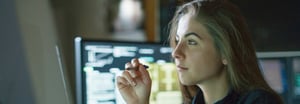 Brunette woman in a dark room reviewing product information on a screen with another monitor behind her.