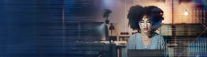 A curly-haired woman in round glasses working on a laptop