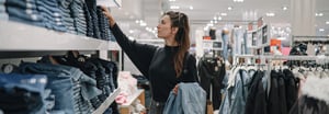A woman browses folded denim on shelves in a clothing retail store, holding a jacket while shopping.
