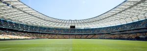 A wide-angle view of a large, modern stadium with a bright green pitch, blue and yellow seating, and a sweeping white roof canopy under a clear sky.