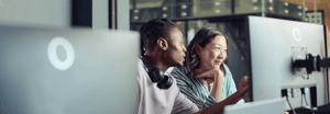 A young man points at a computer monitor, explaining something to his female co-worker