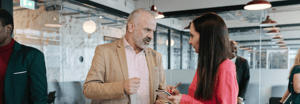 Two professionals engaged in conversation in a modern office, with one taking notes, and colleagues visible in the background.