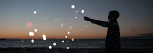 A child's silhouette holding a sparkler on a beach at dusk, with soft bokeh lights scattered across a twilight sky.