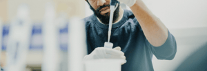 A researcher wearing gloves uses a pipette to transfer a sample into a container in a laboratory setting.