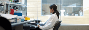 A scientist in a white lab coat working at a desk surrounded by lab supplies, with a large window behind her.