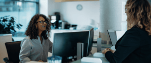 A smiling receptionist behind a service desk having a friendly interaction with a visitor in a bright office setting.
